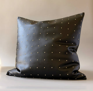 A square black faux-leather throw pillow with small gold studs, set against a plain beige background.