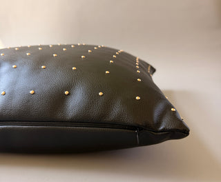 A close-up of a black leather pillow or cushion with small gold studs, photographed on a plain light beige background.