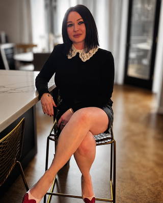 Loren Estes sitting on a gold chair wearing black top with lace collar and black skirt with pink high heels that have a rhinestone heart toe.