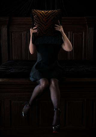 Loren Estes editorial photo with one of her pillows Sable in her bedroom wearing a black dress, hose and high heels